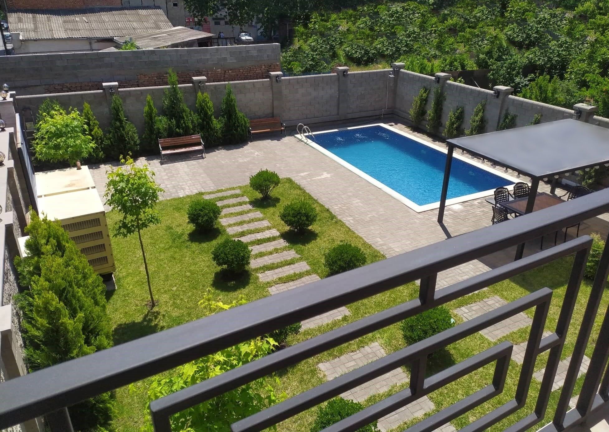 A rooftop pool area with a rectangular pool, surrounded by a low stone wall, a few benches, and small trees.