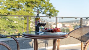 A glass table with a bottle of wine, two glasses, and a plate of food on a balcony with a view of trees and buildings. - Vinum Hotel (Tbilisi)