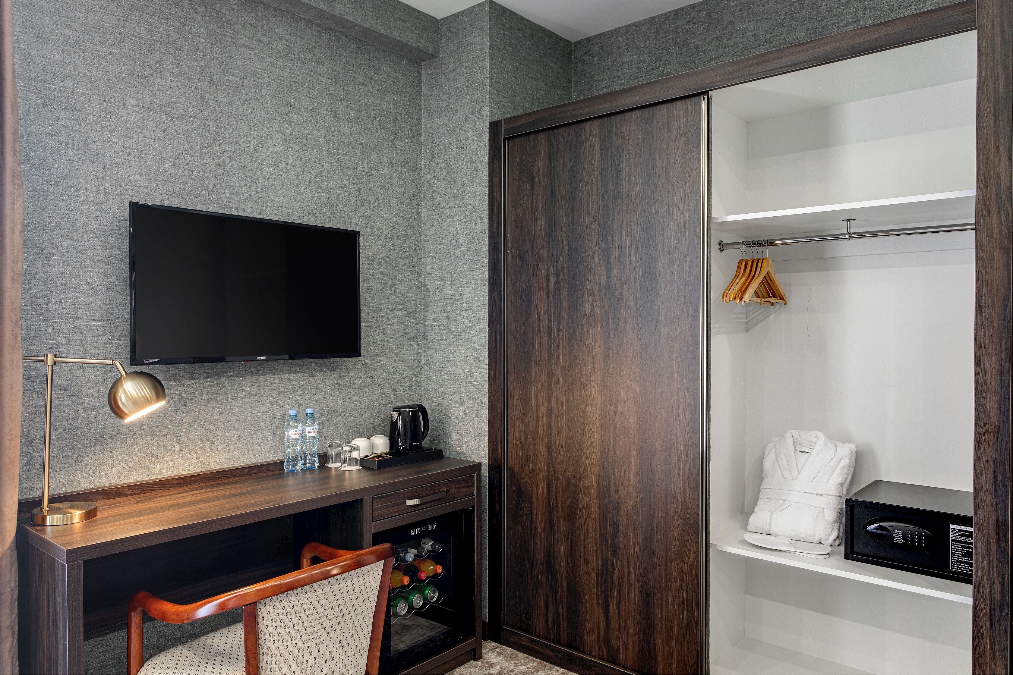A hotel room with a dark wood wardrobe, a flat-screen TV mounted on the wall, a desk with a lamp, a coffee maker, and a minibar.
