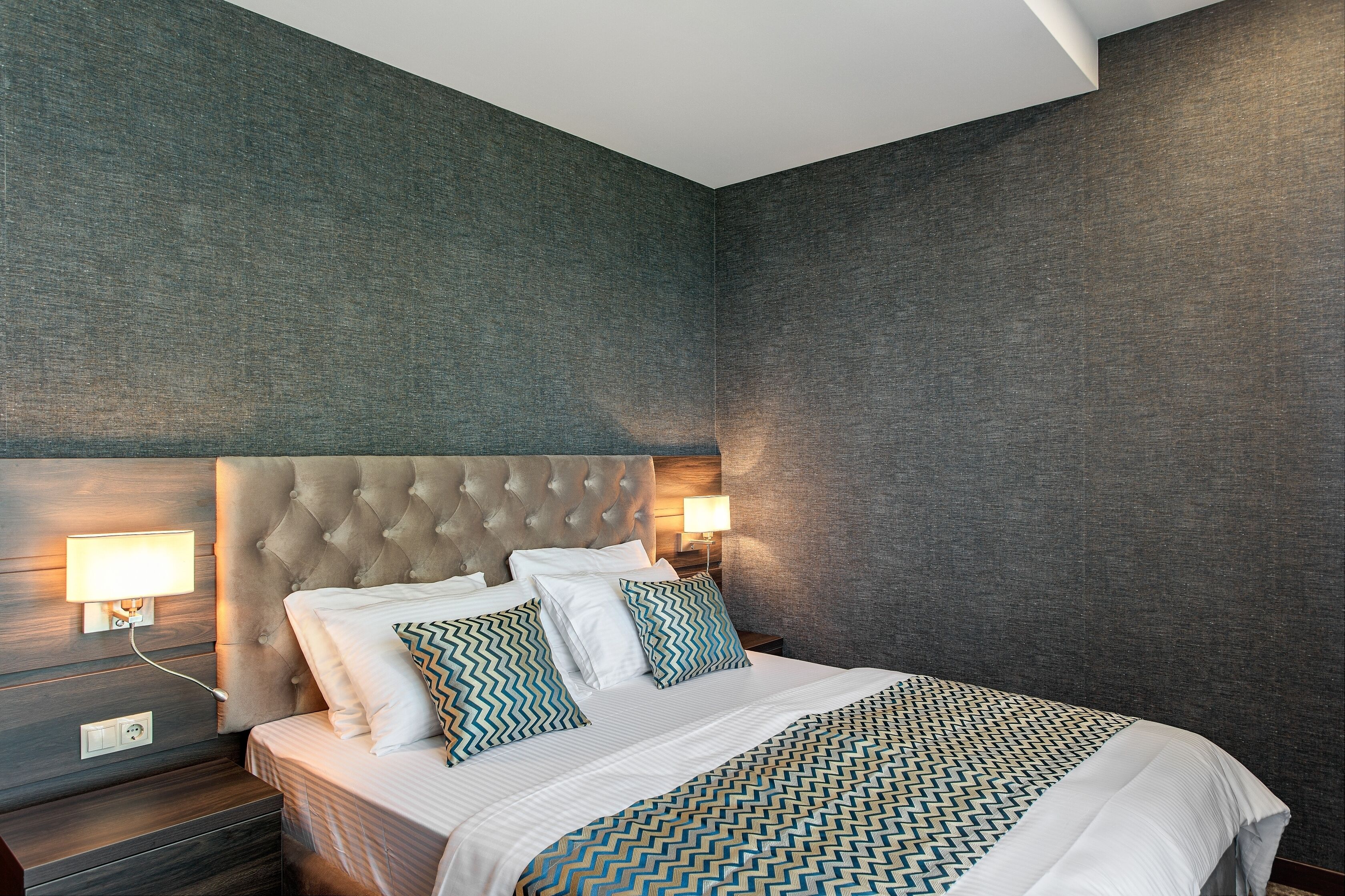 A modern hotel room with a tufted headboard, a large bed with patterned bedding, two bedside lamps, and a dark textured wall.