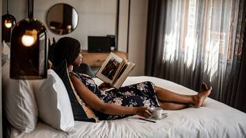 A person lying on a bed reading a book, with a cup and saucer, in a room with a mirror, pendant lights, and a window with curtains.