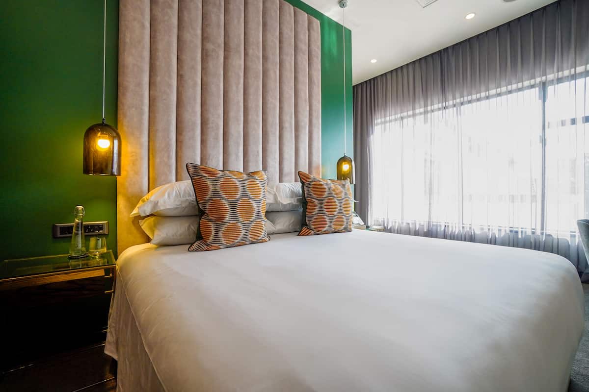 Une chambre d’hôtel avec un grand lit, deux coussins à motifs, une table de chevet surmontée d’une carafe en verre et une vue sur la ville à travers de fines rideaux.