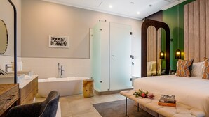 A modern bedroom with a large bed, a bedside table with books, a vanity with a mirror, a bathtub, and a green accent wall.