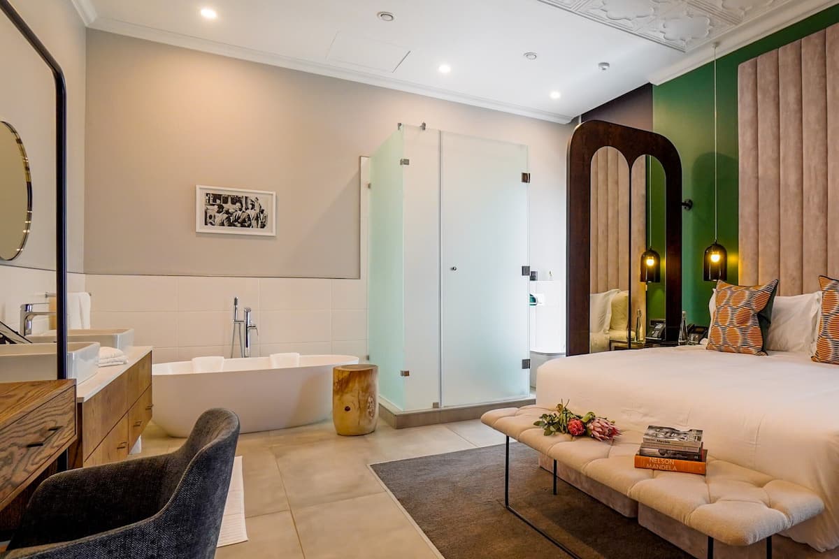 Une chambre moderne avec un grand lit, une table de chevet avec des livres, une coiffeuse avec un miroir, une baignoire et un mur d’accent vert.