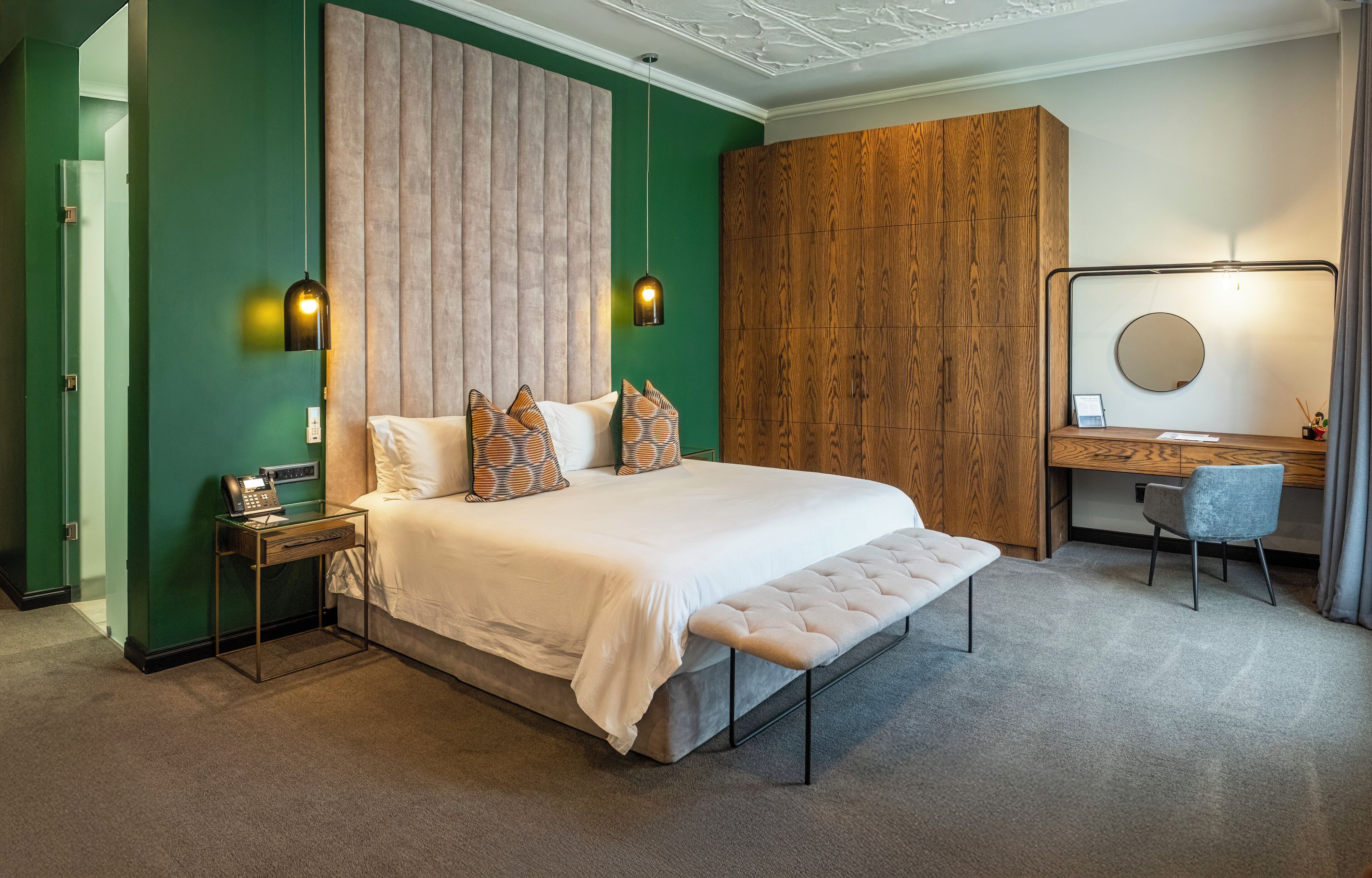 A modern hotel room with a large bed, a wooden wardrobe, a desk with a chair, and a mirror.