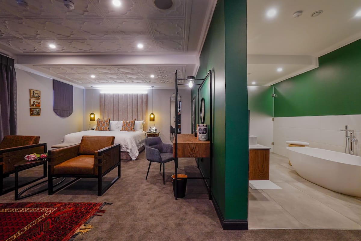 Une chambre d’hôtel comprenant un lit, deux fauteuils, une petite table, un bureau, un miroir, un mur d’accent vert et une salle de bain avec une baignoire.