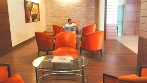 Eine Hotelhalle mit orangefarbenen Stühlen, einem Glastisch und einer Steinwand.