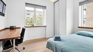 Une chambre moderne avec un bureau en bois, un fauteuil de bureau noir, un lit avec une literie bleue et une fenêtre donnant sur des immeubles et des arbres.