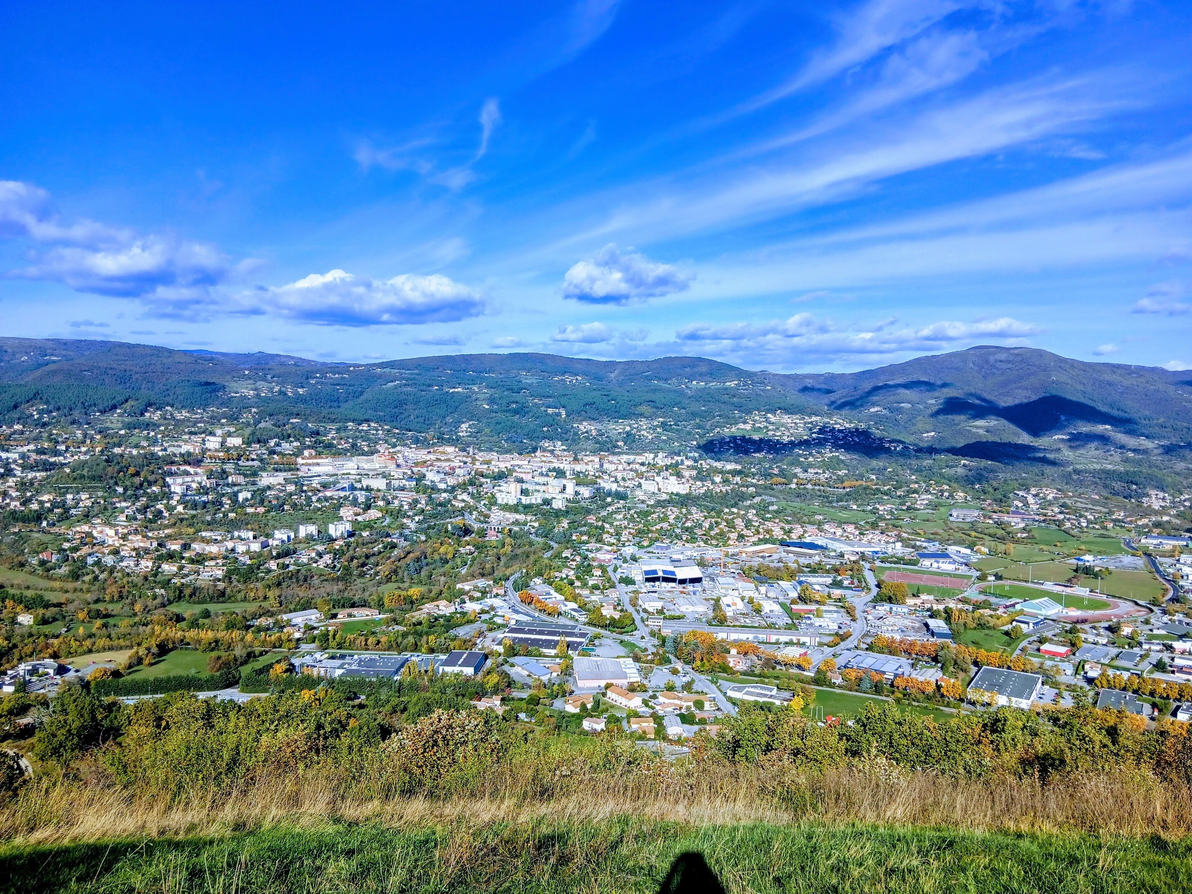 A panoramic view of a town with a mix of residential and commercial buildings, surrounded by hills and greenery.