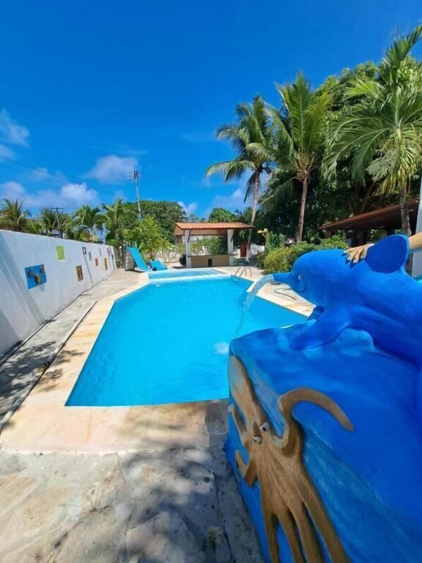 A swimming pool with a blue water feature, surrounded by palm trees and a white wall. - Pousada Neptun (Conde)