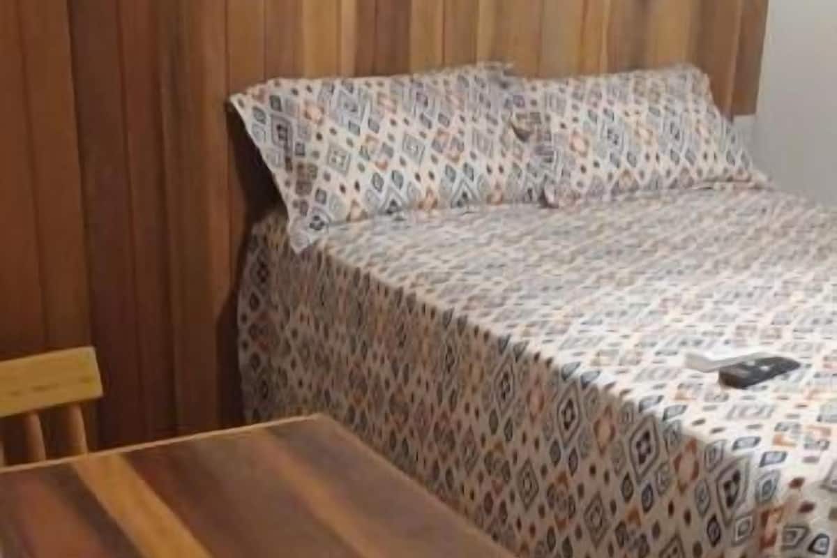 A room with a wooden bed, a patterned bedspread, a wooden table, and a chair.