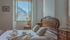 A bedroom with a bed, a wooden nightstand, a chair, a window with a mountain view, and a framed picture on the wall. - Casa Ferrari in Varenna (Varenna)