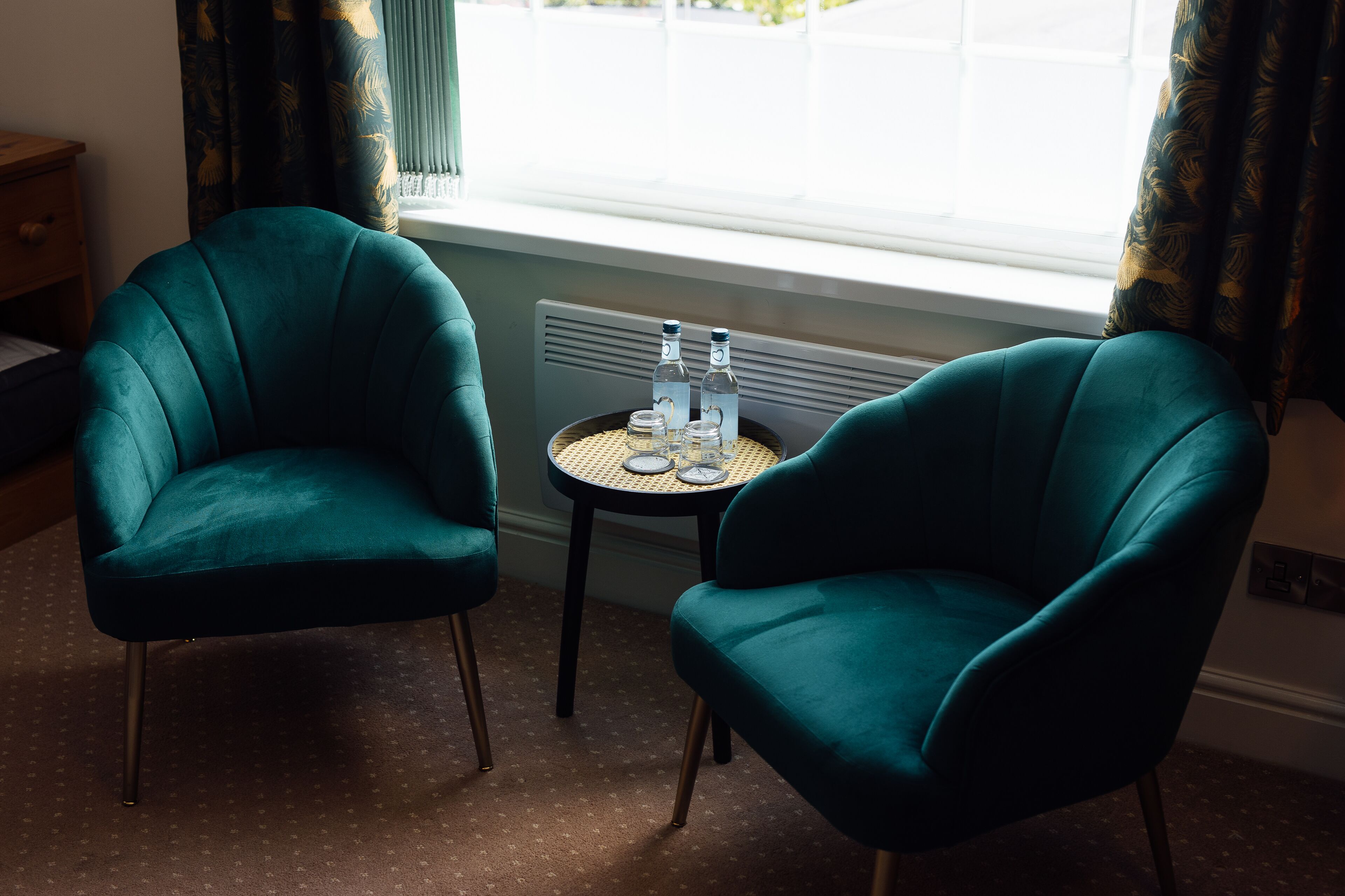 Two teal armchairs facing each other with a small round table between them, containing two bottles and glasses.