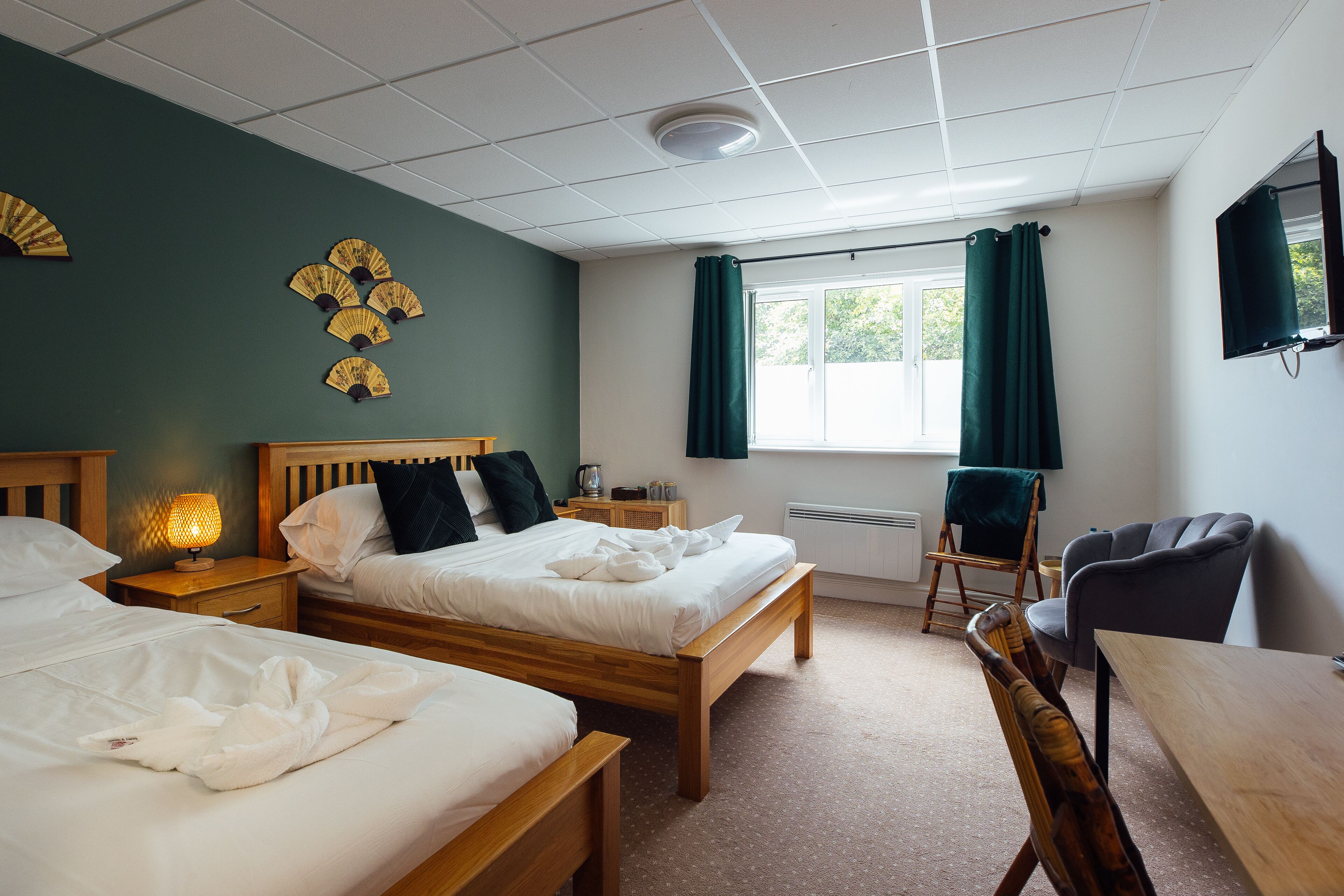 A hotel room with two beds, a desk, a chair, a window with curtains, a wall-mounted TV, and a decorative fan on the wall.