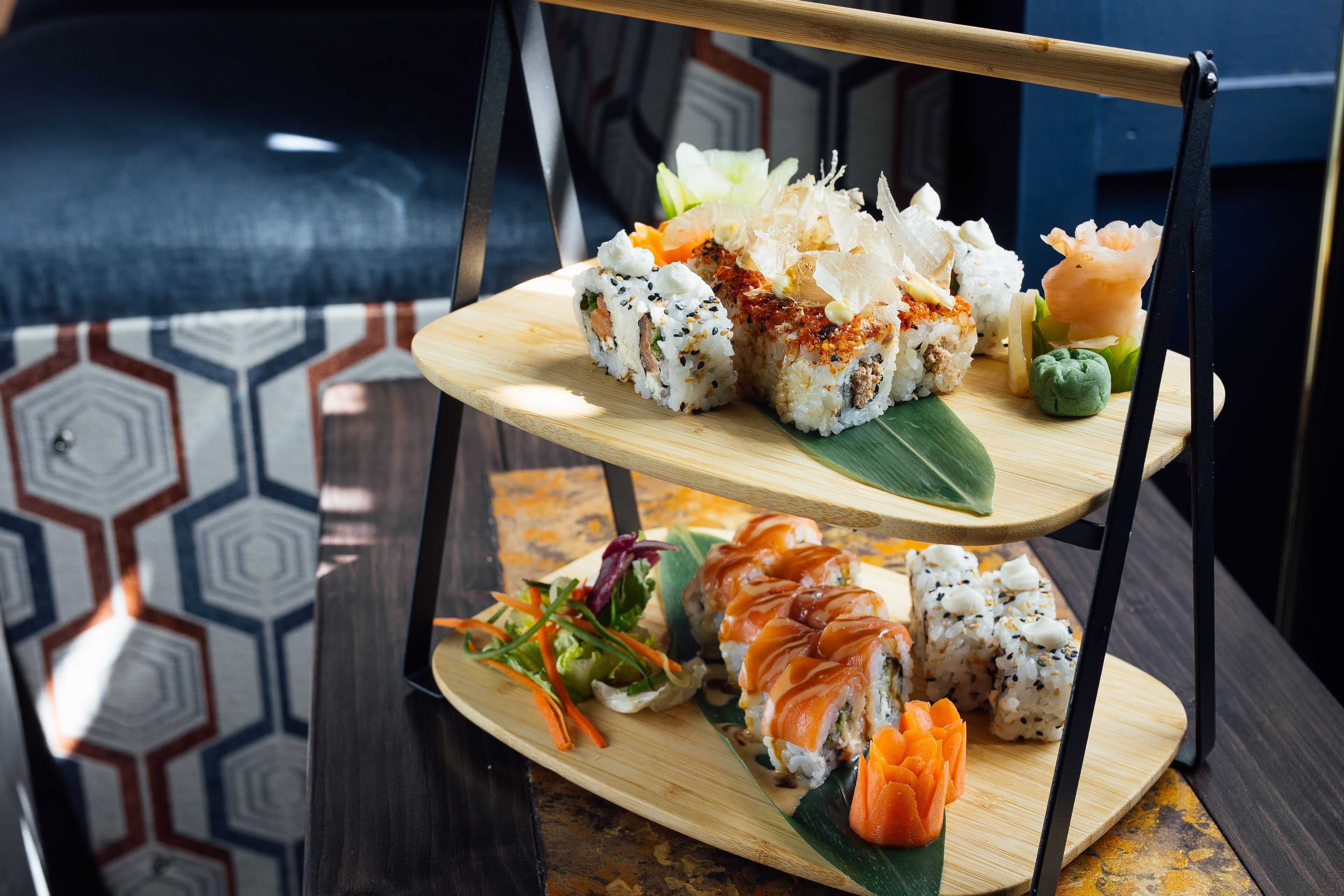 A multi-tiered wooden stand displaying various sushi dishes, including rolls and sashimi, on a table with a geometric patterned floor.