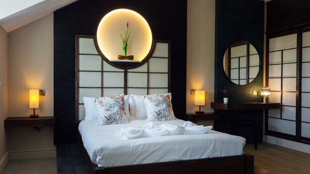 A modern hotel room with a large bed, bedside lamps, a round mirror, and a wall-mounted shelf with a plant. - The Imperial Dragon Hotel (Saundersfoot)