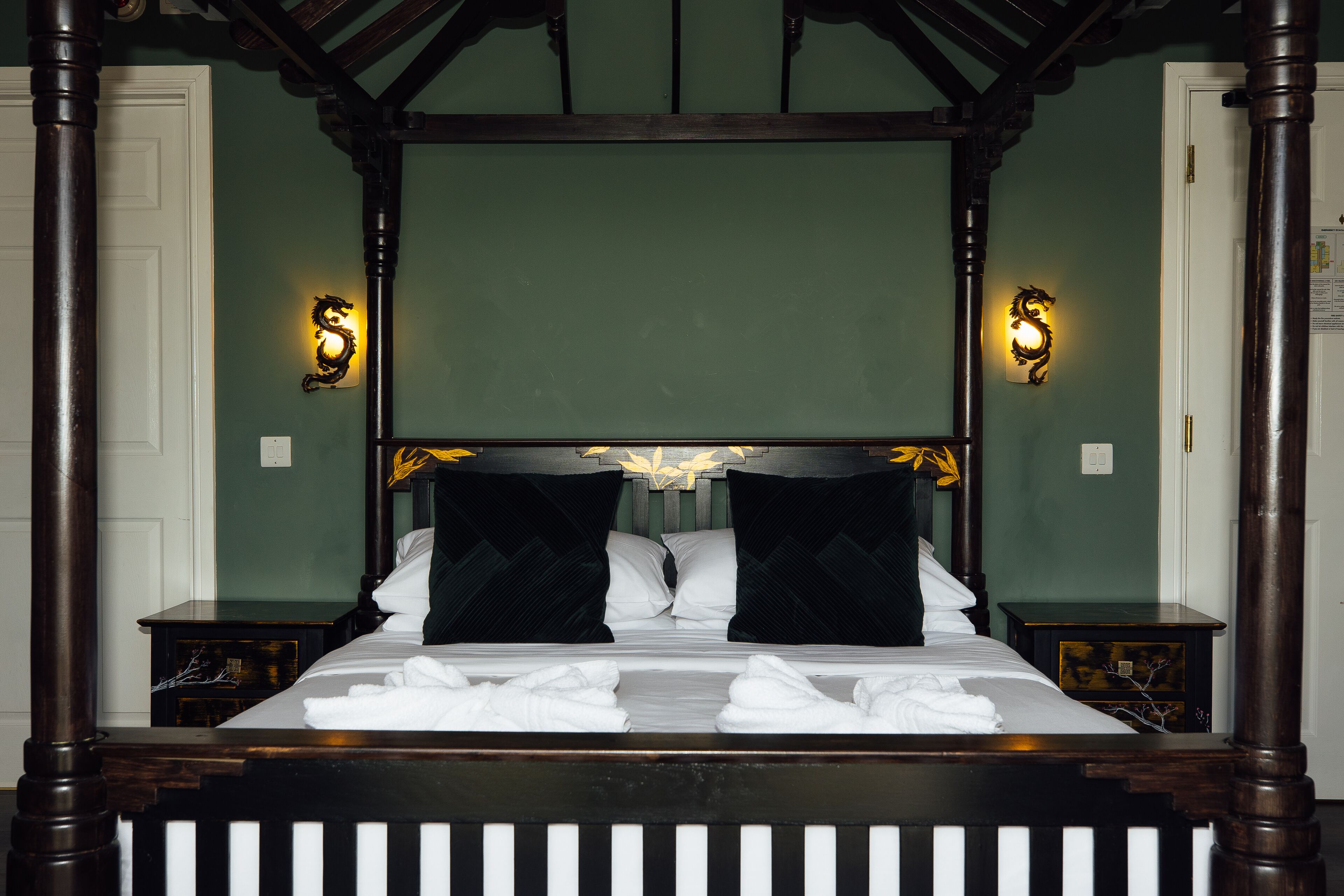 Una cama con dosel, ropa de cama blanca y almohadas negras, dos mesitas de noche y apliques con un diseño decorativo.