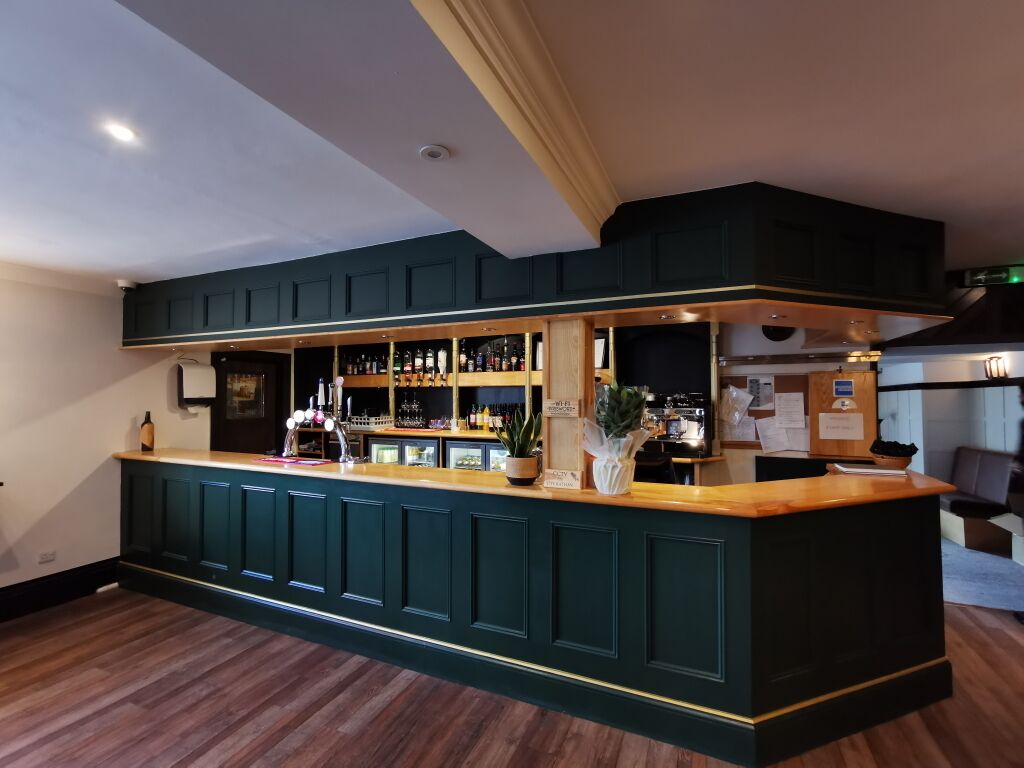 A bar area with a dark green counter, wooden flooring, and a ceiling with recessed lighting.