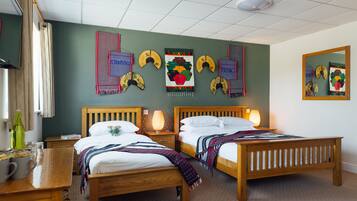 A hotel room with two wooden beds, a nightstand with a lamp, a wooden dresser, and a wall decorated with fans and textiles.