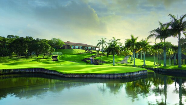 Een golfbaan met een waterpartij, palmbomen en een gebouw op de achtergrond.