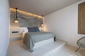 Une chambre moderne avec un mur d’accent en pierre, un lit recouvert d’une couverture grise, deux lampes suspendues et un fauteuil.