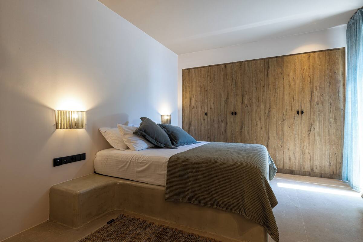 A modern bedroom with a bed, bedside lamps, a wooden wardrobe, and a view of the outdoors.