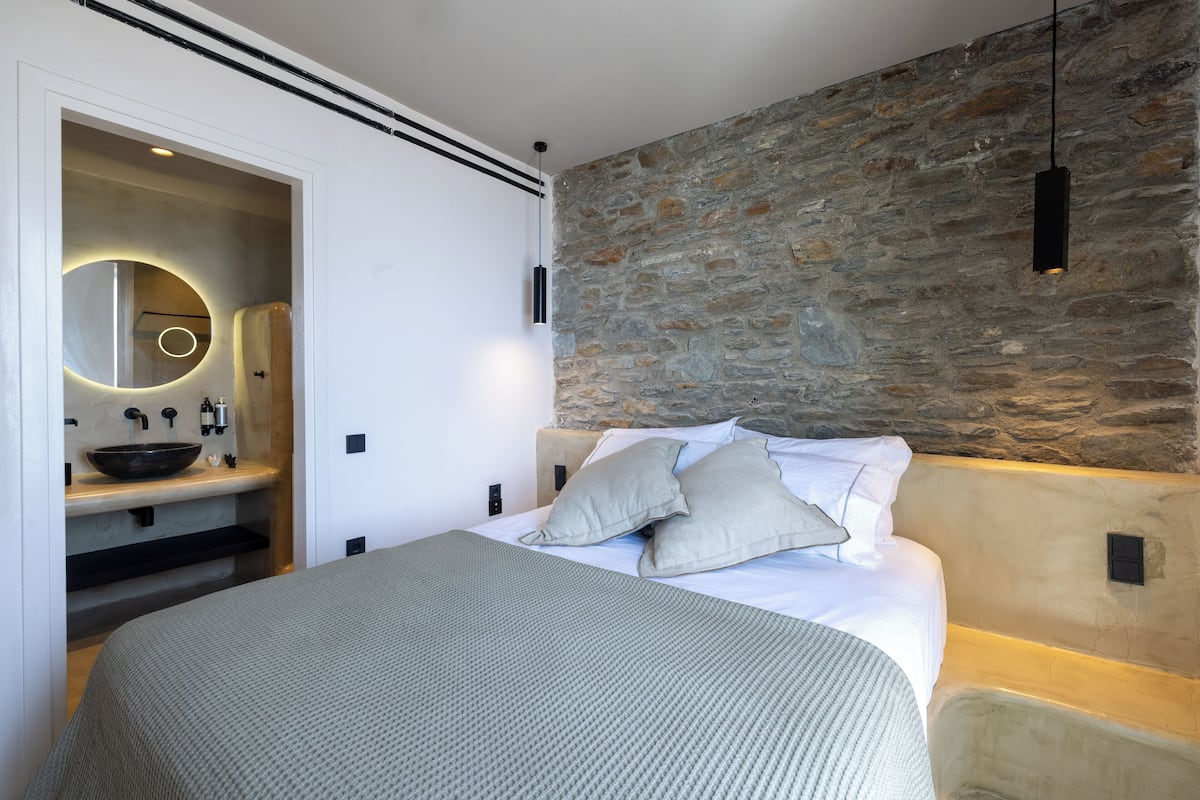 A modern bedroom with a stone accent wall, a round mirror, a built-in sink, and a large bed with pillows.