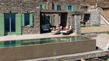 A stone pool with lounge chairs, a stone building with green shutters, and a hillside in the background.