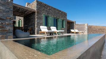 A modern villa with a stone exterior, a swimming pool, and two lounge chairs.