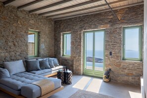 A stone-walled room with a sofa, a small table, and a view of the sea through large windows.