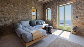A stone-walled room with a modern sofa, a small side table, and a view of the sea through a green-framed window. - Kalma Living - Luxury Villas (Kithnos)