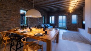 A spacious dining area with a wooden table set for four, stone walls, and a large hanging light fixture. - Kalma Living - Luxury Villas (Kithnos)