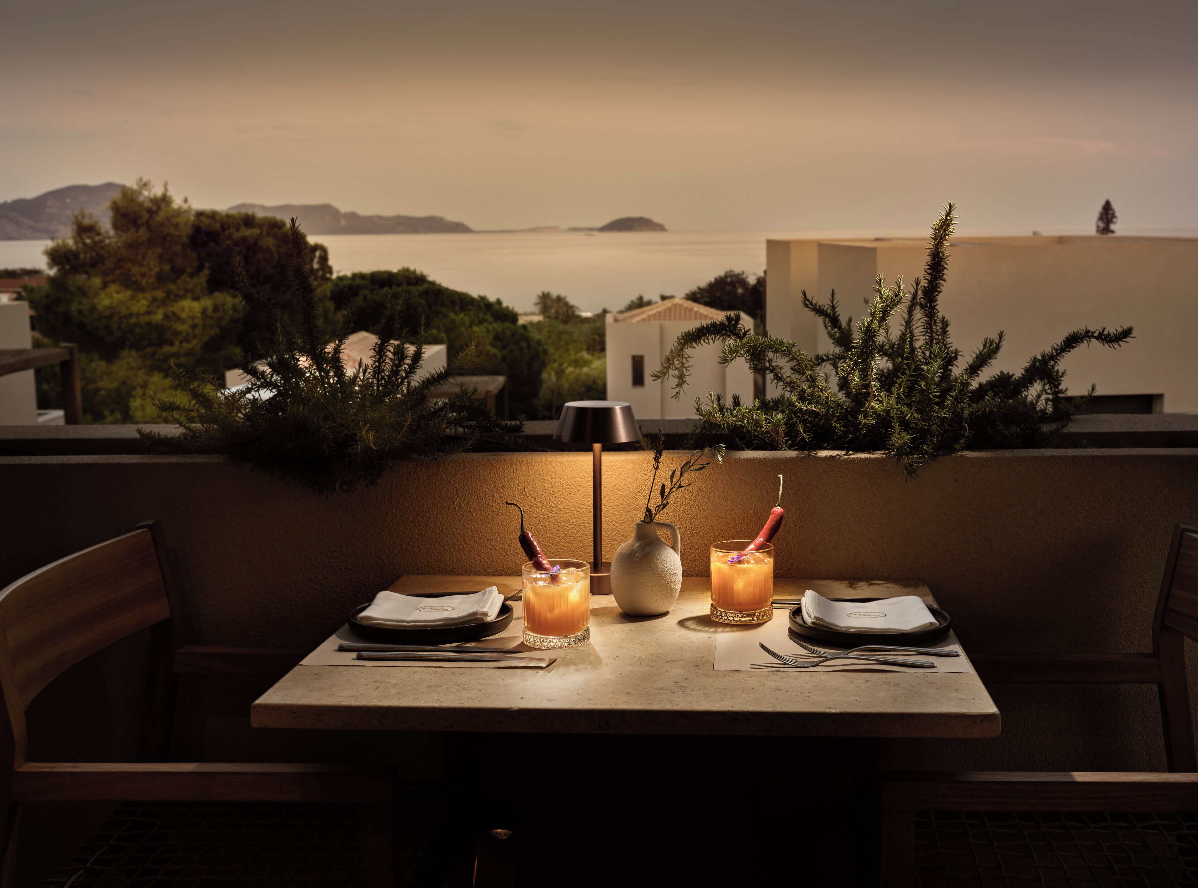 Mesa para dos con bebidas, un jarrón y una lámpara, con vistas a la costa, edificios y vegetación.