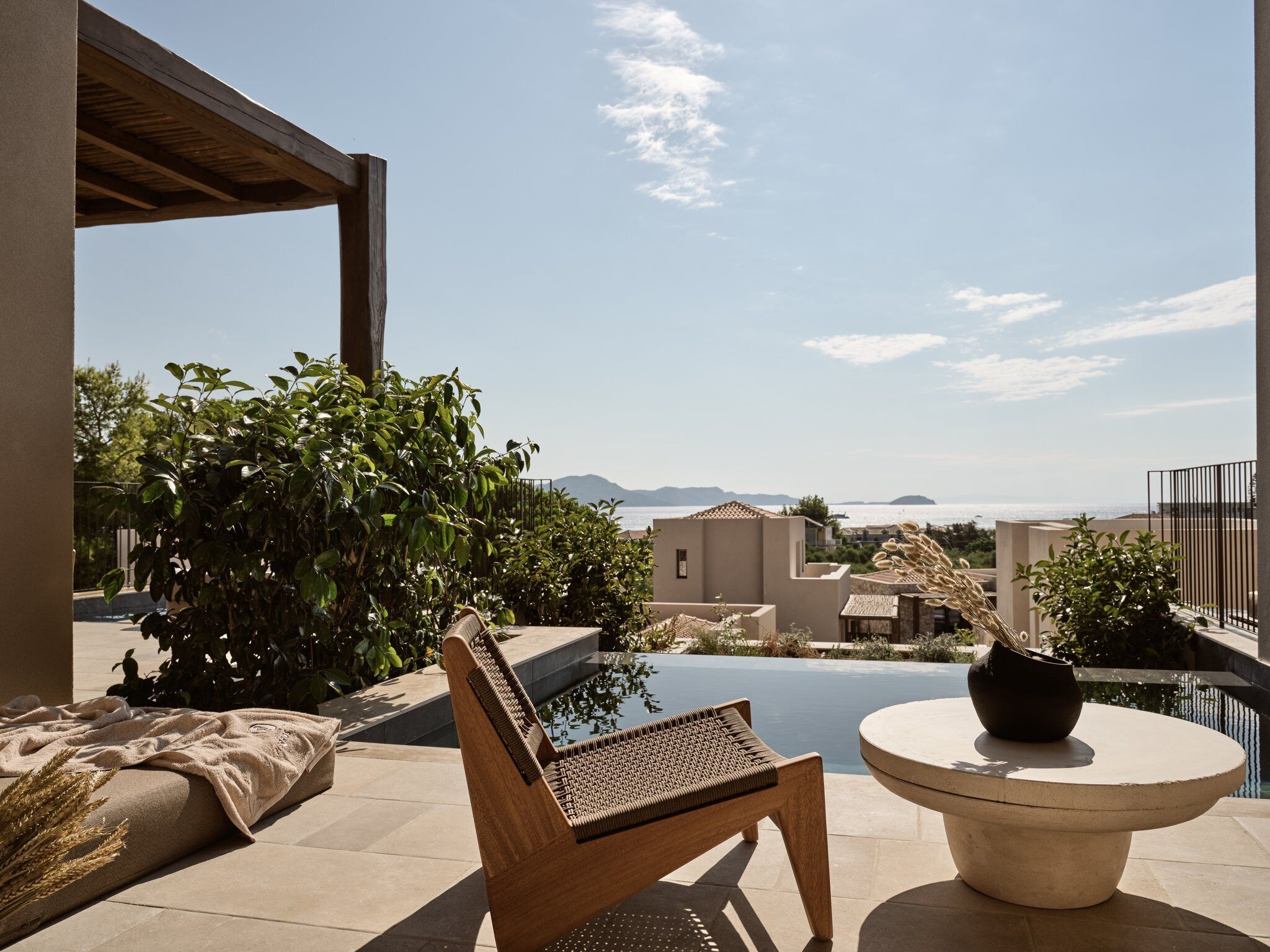 Un área de estar al aire libre con piscina, mobiliario de madera y vistas al mar y a las montañas.