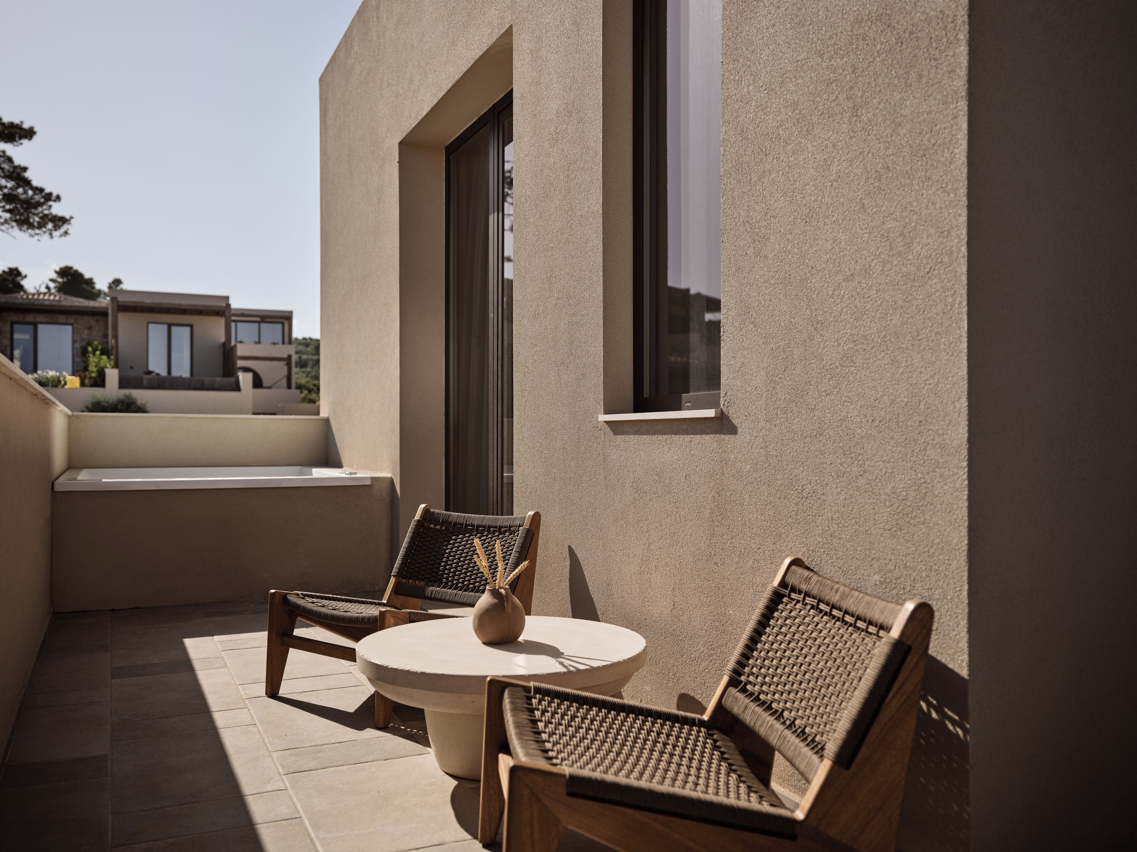 A patio with two wicker chairs and a round table, a small vase, and a window.