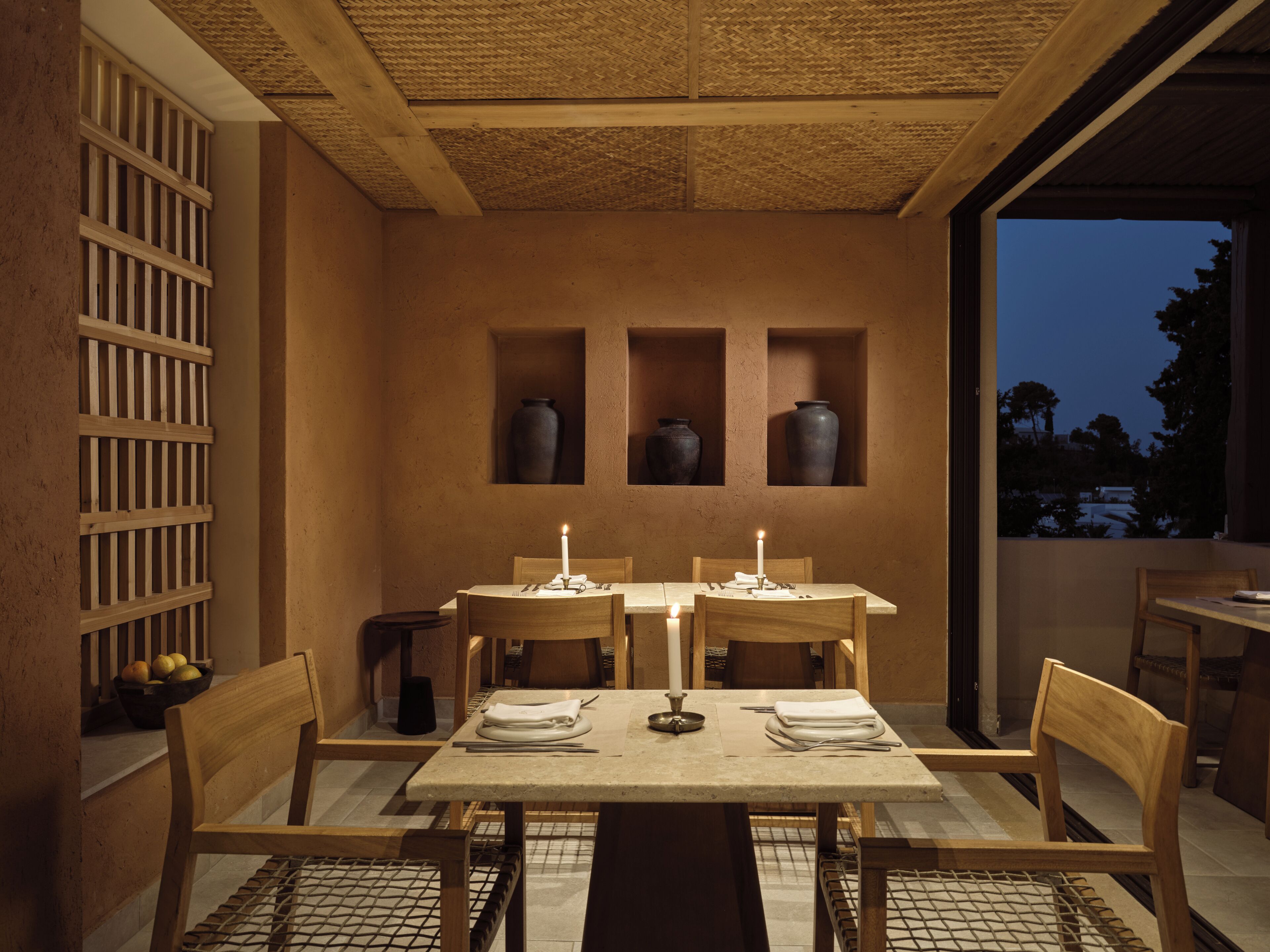 A dining area with a table set for two, wooden chairs, and a view of the outdoors at dusk.