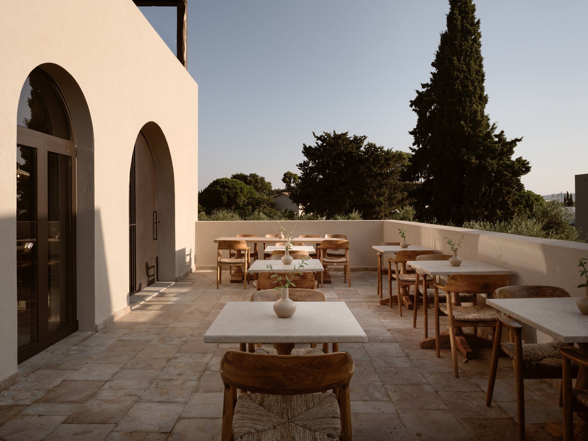 An outdoor patio with multiple tables and chairs, a stone floor, and a view of trees and buildings in the background.
