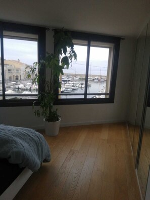 Een kamer met een groot raam dat uitkijkt op een jachthaven, een bed en een potplant.