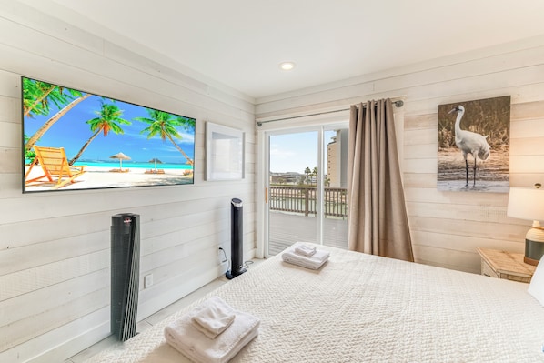 A hotel room with a bed, a TV displaying a beach scene, a standing heater, and a balcony with a view of the beach.