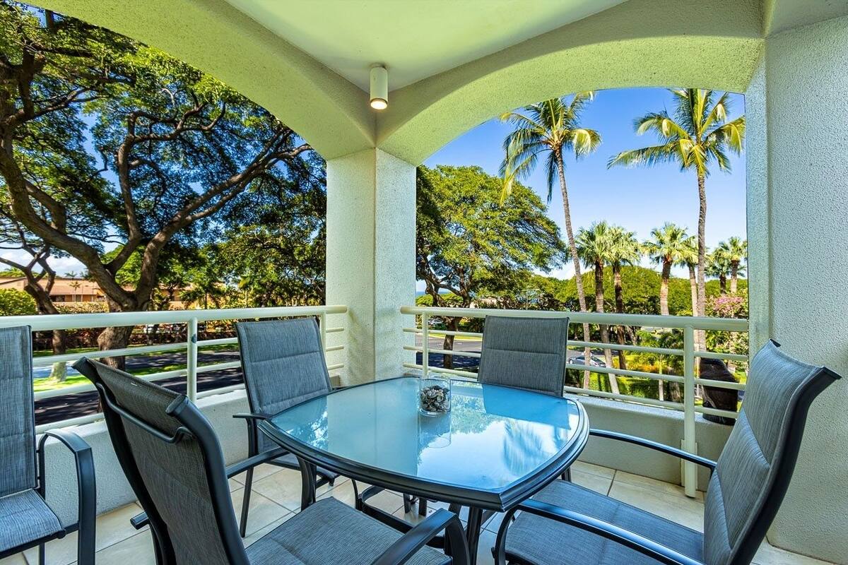 A glass-top table with four chairs, a white railing, and a view of palm trees and a road.