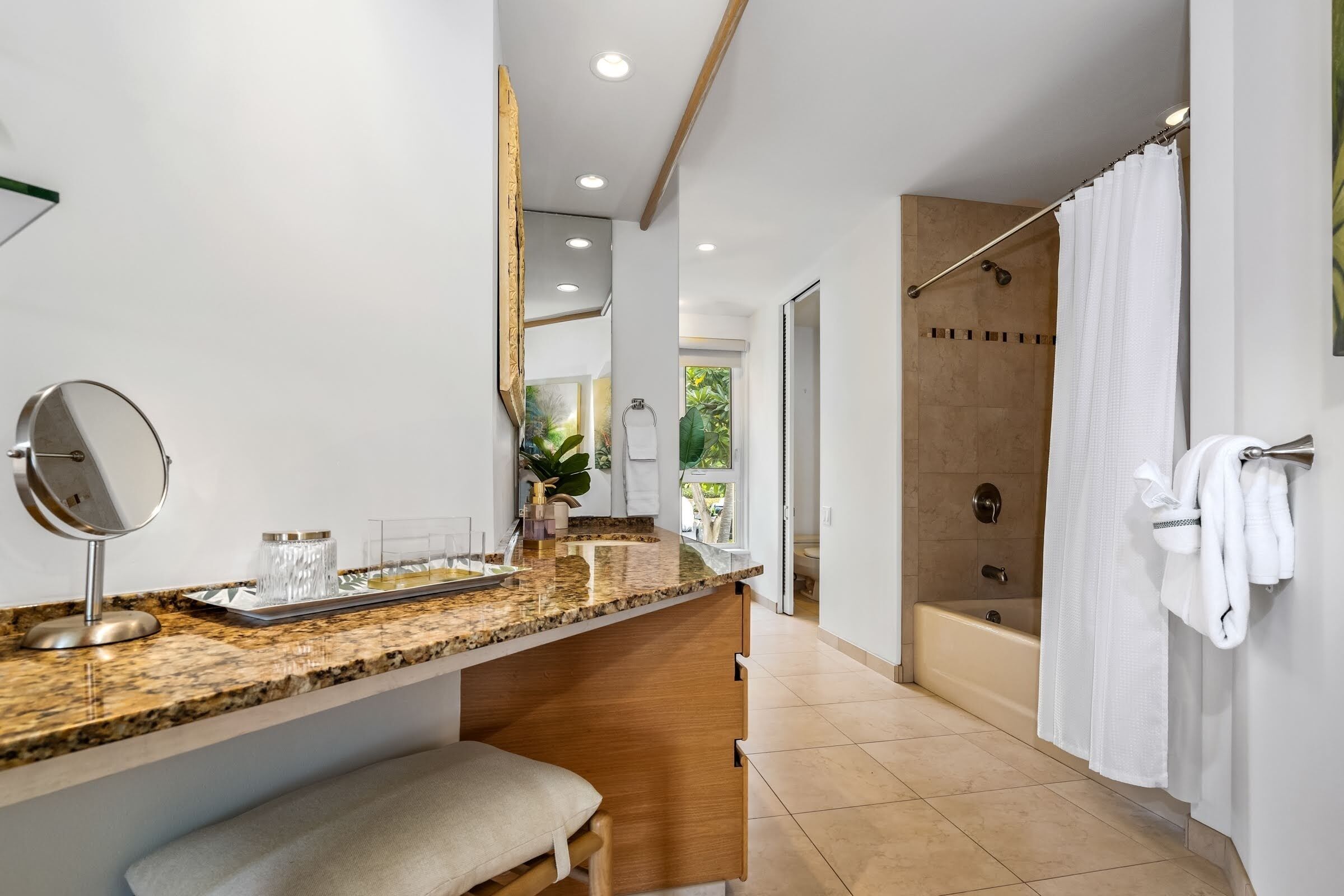 A modern bathroom with a granite countertop, a bench, a shower with a glass door, and a large mirror.