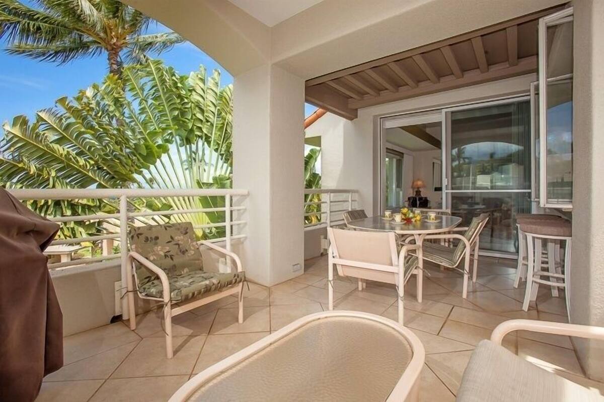 A balcony with white wicker furniture, a glass door leading to a dining area, and tropical plants.