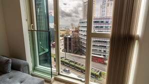 A modern room with a large window overlooking a city street, featuring a glass partition, a grey sofa, and a view of urban buildings and a tram. - Luxury 2-bed Croydon Apartment Near Gatwick (Croydon)