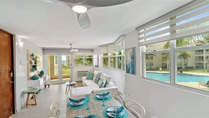 Un salon spacieux avec un canapé blanc, une table à manger dressée pour quatre personnes et une grande fenêtre donnant sur une piscine.