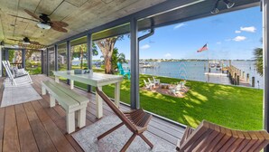 A covered outdoor area with wooden flooring, white benches, and a table. It features large windows offering views of a marina and a flag.