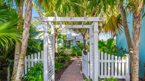 A pathway leading through a white wooden gate into a lush garden area with palm trees and a blue building in the background.