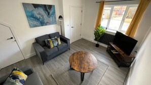 A living room with a sofa, a coffee table, a TV, and a painting on the wall. - Brand New High Spec House with parking (Newcastle-Under-Lyme)