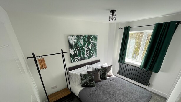 A bedroom with a bed, a radiator, a window with curtains, and a wall-mounted light fixture. - Brand New High Spec House with parking (Newcastle-Under-Lyme)