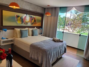 A modern hotel room with a large bed, a painting above the bed, a view of the outdoors, and a nightstand with a lamp and a bottle of wine. - Villa Terragona (Alto Paraíso de Goiás)