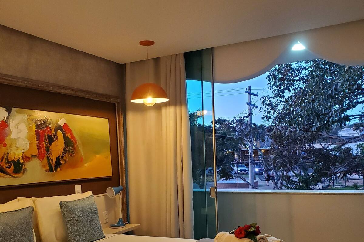 A hotel room with a large bed, a painting on the wall, a window with a view of the outside, and a nightstand with a lamp.